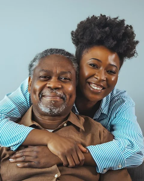 An adult daughter sits close beside her elderly father on the sofa at home, her arms around him, both at ease.