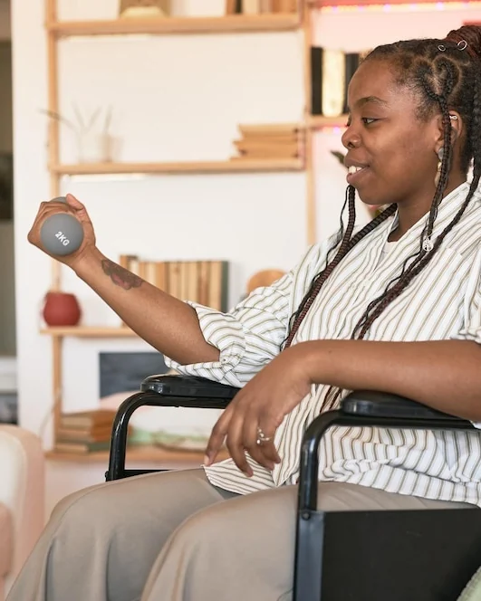 A woman in a wheelchair lifting a small dumbbell during a strength session at home.